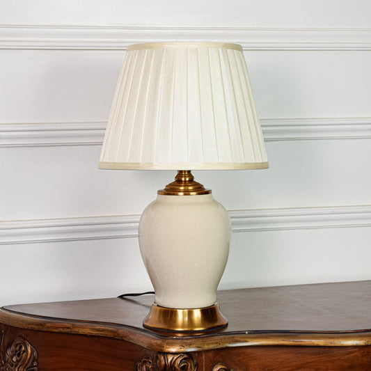 Classic ivory ceramic table lamp with gold metal base and pleated fabric shade placed on soft off white background.