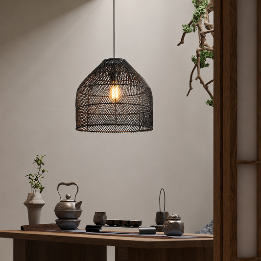 A black woven rattan pendant light hangs over a wooden table in a styled interior setting with tea accessories and plants.