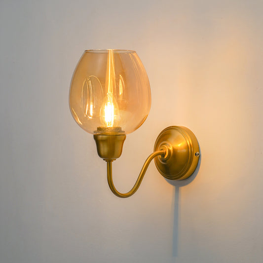 A wall-mounted sconce with a warm brass/gold metal arm and circular backplate, featuring a clear glass globe shade that glows with an exposed filament bulb against a plain light wall.