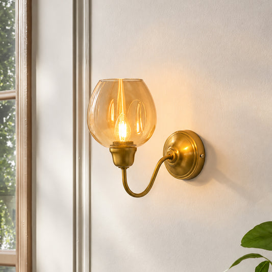 A warm brass wall sconce with a clear glass globe shade, mounted on a white wall next to a window.