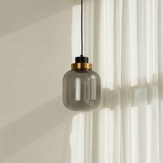 A modern pendant light with a smoky translucent glass shade, a black top cap, and a brushed brass ring, hanging against a warm beige wall with white sheer curtains.