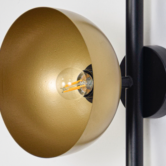 Wall light closeup highlighting smooth gold metal shade with glowing bulb and slim black bar showcasing premium craftsmanship and sculptural design.