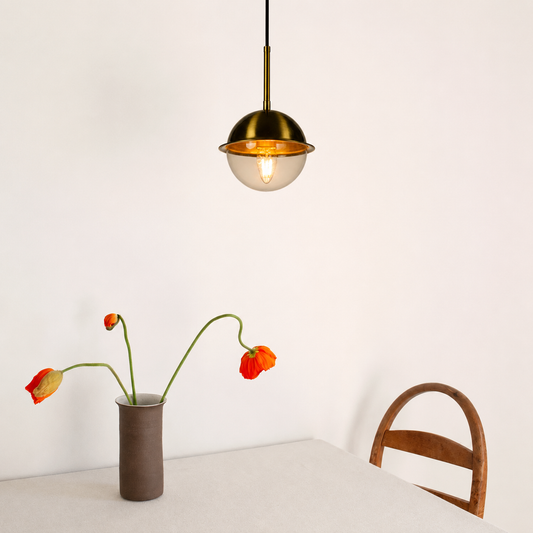 A modern pendant light with a brass-finished dome shade and a clear glass lower bowl, hanging above a table in a minimalist room. A brown vase with orange flowers and part of a wooden chair are visible below.