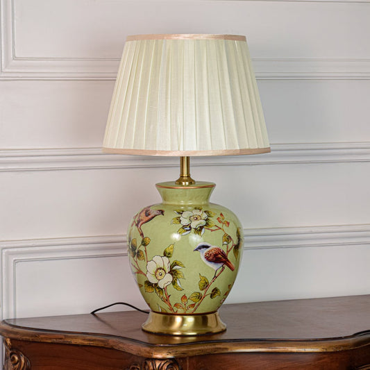 Decorative green ceramic table lamp with bird and floral design brass base and pleated fabric shade on off white background.