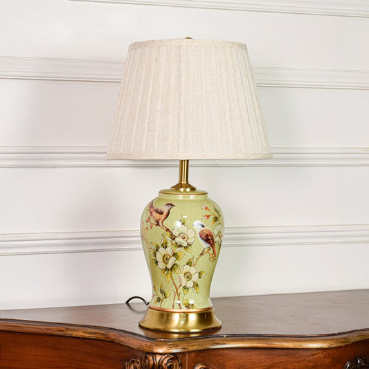 Decorative green ceramic table lamp with bird and floral design brass base and fabric shade on soft off white background.
