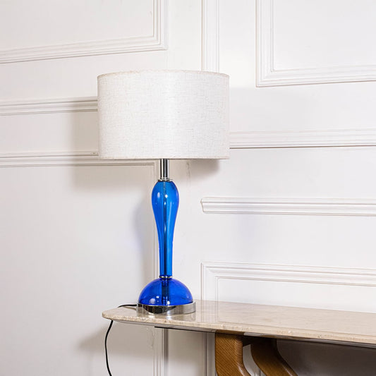 Blue glass lamp with lights off highlighting glossy glass body and fabric shade on plain white background.