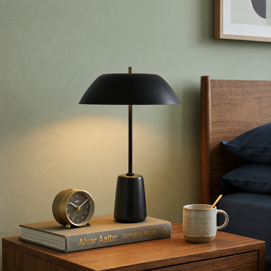 A modern black table lamp with a conical shade and cylindrical base sits on a wooden nightstand beside a book, a vintage-style alarm clock, and a ceramic mug in a bedroom setting.