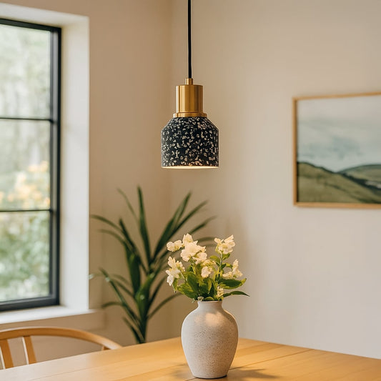 Black terrazzo pendant light with gold metal top glowing warmly above a wooden table with a ceramic vase and white flowers in a modern dining space.