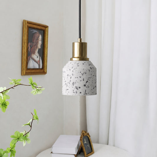A modern pendant light with a white terrazzo-patterned glass shade and a brushed brass metal collar, hanging in a bright, minimalist room with white curtains, a framed picture, and a small table below.