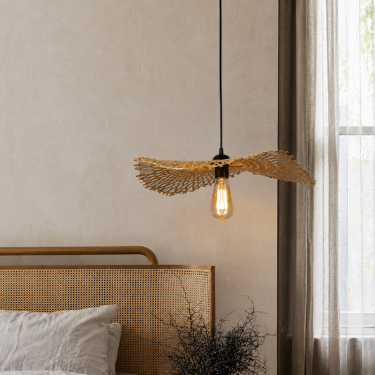 A hanging pendant light with a woven natural-fiber bird-shaped shade and a glowing Edison bulb, shown above a bed in a softly lit bedroom.