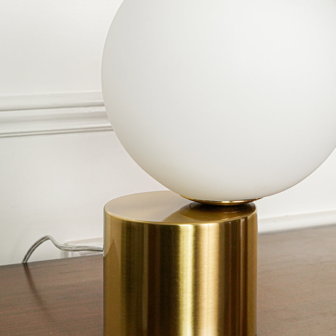 Minimal white globe table lamp with metallic gold base positioned on wooden surface against soft neutral wall background ideal for modern decor.