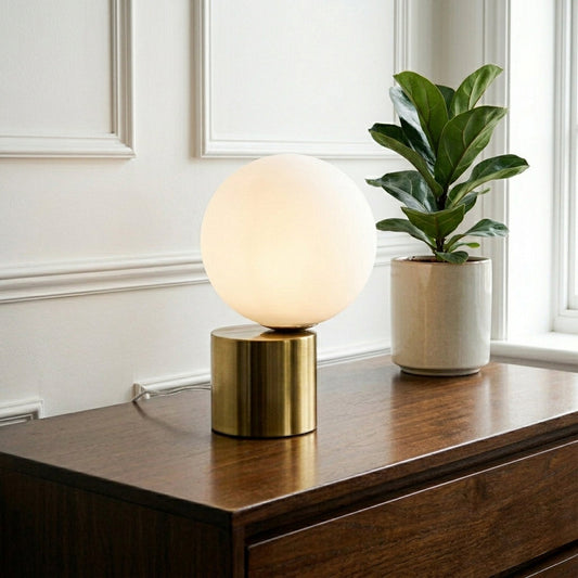 Modern white globe table lamp with gold metal base placed on wooden console against textured white wall background ideal for luxury home decor.