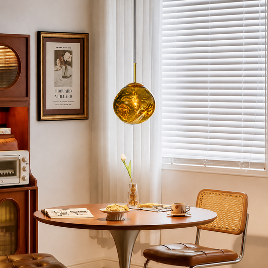 A round wooden dining table in a cozy room setting, styled with a brown leather and cane chair, a hanging amber glass pendant light, and a framed print on the wall.