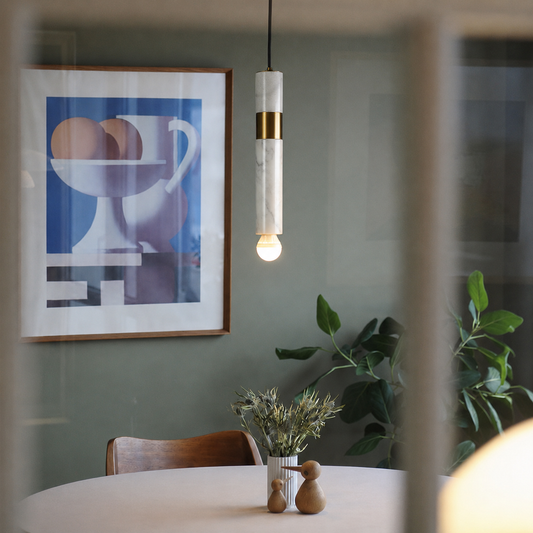 A modern cylindrical pendant light with a white marble-look body and gold band hangs over a dining table in a real interior setting, with framed wall art, a chair, and a plant visible in the background.