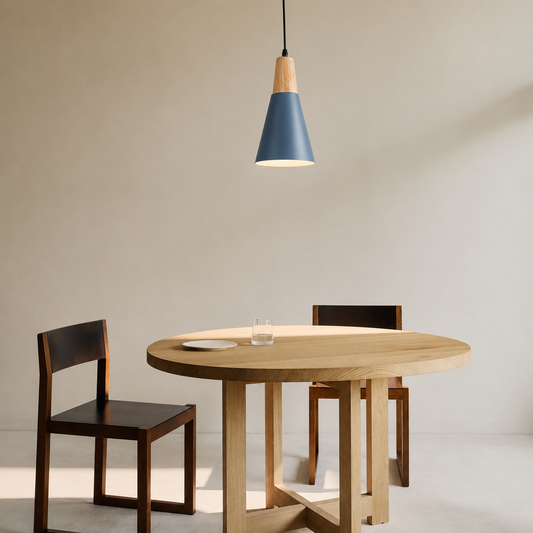 A modern dining setup featuring a round light oak table, two dark wood chairs with black seats, and a blue and wood conical pendant light hanging above against a neutral beige wall.