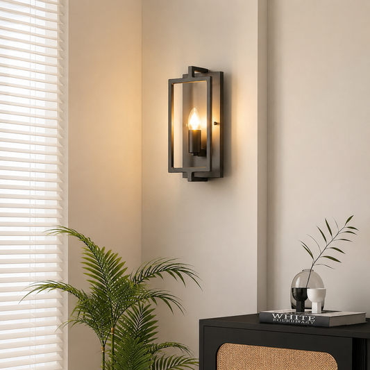 A modern rectangular wall sconce with a dark metal frame and glowing light bulb, mounted on a beige wall in a styled interior next to a window with blinds and a potted plant.
