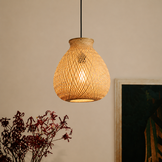 A woven rattan pendant light glowing with warm light, hanging in a home setting next to a framed picture and dried plants.
