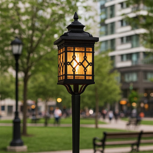 Black metal garden spike light with decorative patterned glass glowing warmly in a park pathway creating an inviting illuminated outdoor atmosphere.