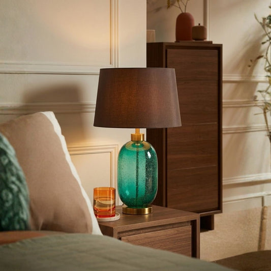A table lamp with a dark brown drum shade and a textured teal-green glass base with gold metal accents, styled on a wooden bedside table in a warm, modern bedroom setting.