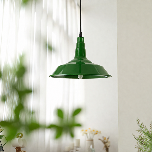 A green metal pendant light with a wide conical shade hanging from a black cord in a softly lit interior setting with blurred plants and decor in the background.