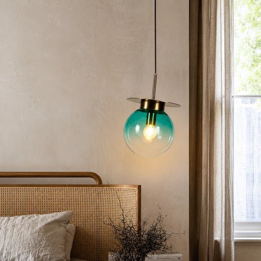 A modern pendant light with a translucent teal glass globe and brass hardware, hanging in a bedroom next to a window.