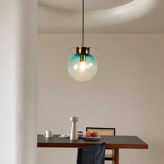 A modern pendant light with a translucent teal-blue glass globe shade and a brass-colored metal top, hanging above a dark wood dining table in a minimalist room.
