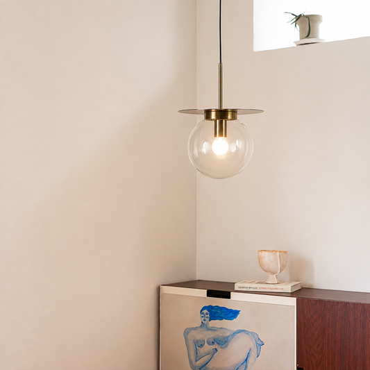 A modern pendant light with a clear glass globe shade and a brass-colored circular canopy, hanging in a minimalist room corner above a wooden cabinet.