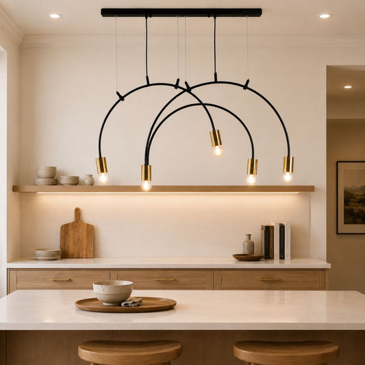 Modern black arched pendant chandelier with five gold-finished cylindrical shades hanging over a kitchen island above a white countertop and light wood cabinetry.