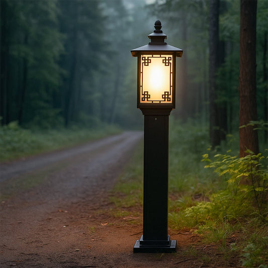 A tall outdoor pathway lamp with a decorative geometric frame glowing warmly in a forest setting casting soft light along a natural dirt trail surrounded by dense green trees at dusk.