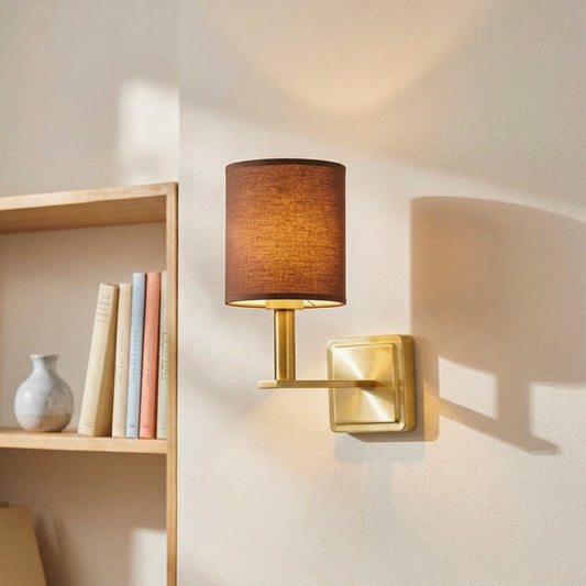 Modern gold metal wall sconce with a dark brown fabric cylinder shade casting warm light beside a wooden bookshelf.