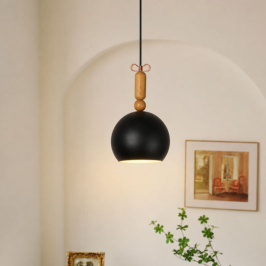 A modern black spherical pendant light hanging from a black cord with a light wood accent and small bow detail, shown in a warm indoor setting against a cream wall with an arched niche, framed art, and a green plant.