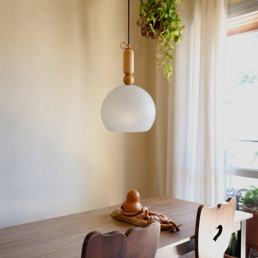 A modern pendant light with a frosted white globe shade and natural wood accents hangs over a wooden dining table in a softly lit room with a hanging plant and window curtains.