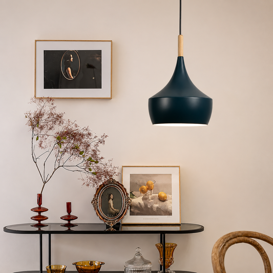 A modern pendant light with a matte dark teal-blue conical shade and a light wood accent near the top, hanging in a styled interior above a black console table with framed art and decorative objects.