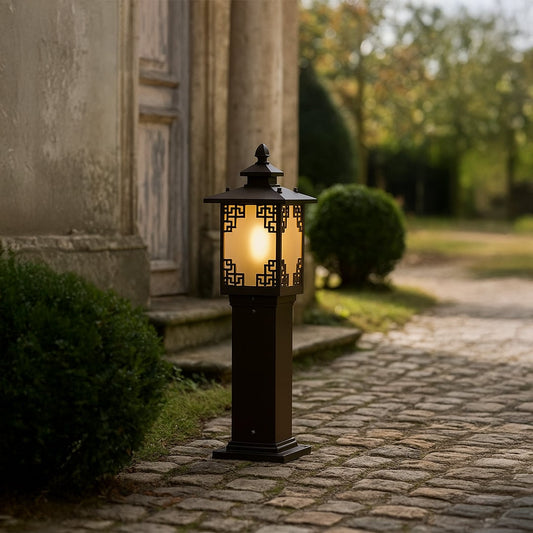 A warm outdoor pathway scene featuring a tall black garden bollard light with decorative panels glowing softly among cobblestone and greenery creating an inviting entrance at dusk.