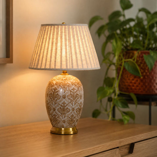A table lamp with a patterned ceramic base and a pleated fabric shade, illuminated on a wooden console table with a blurred potted plant in the background.