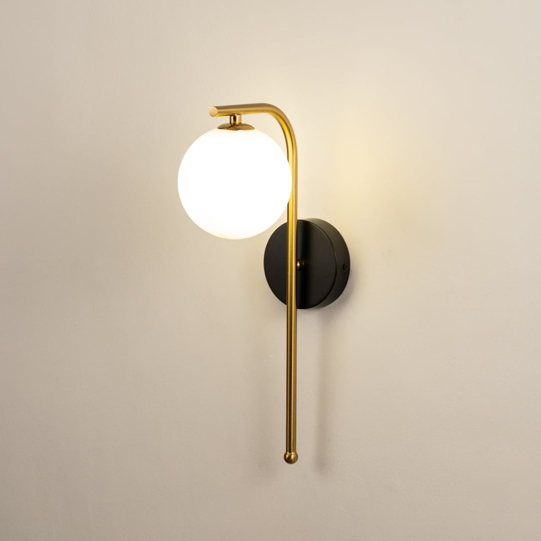A gold and black metal wall light with a frosted globe shade shown with lights on against a white background highlighting its warm ambient glow.