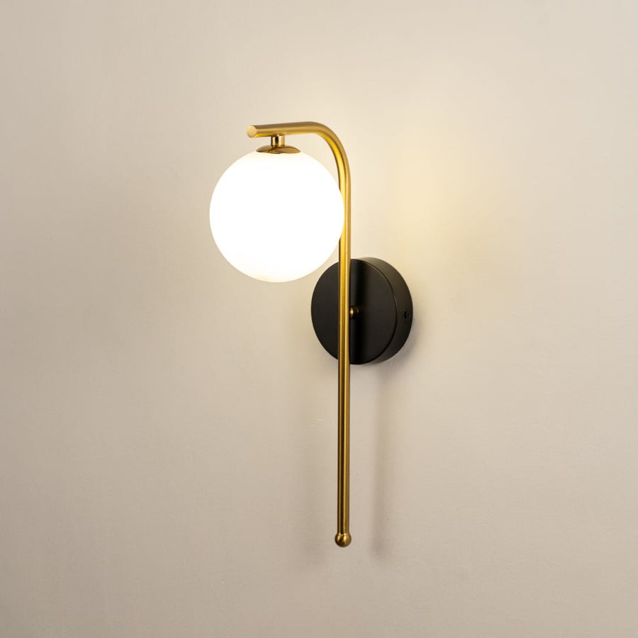 A gold and black metal wall light with a frosted globe shade shown with lights on against a white background highlighting its warm ambient glow.