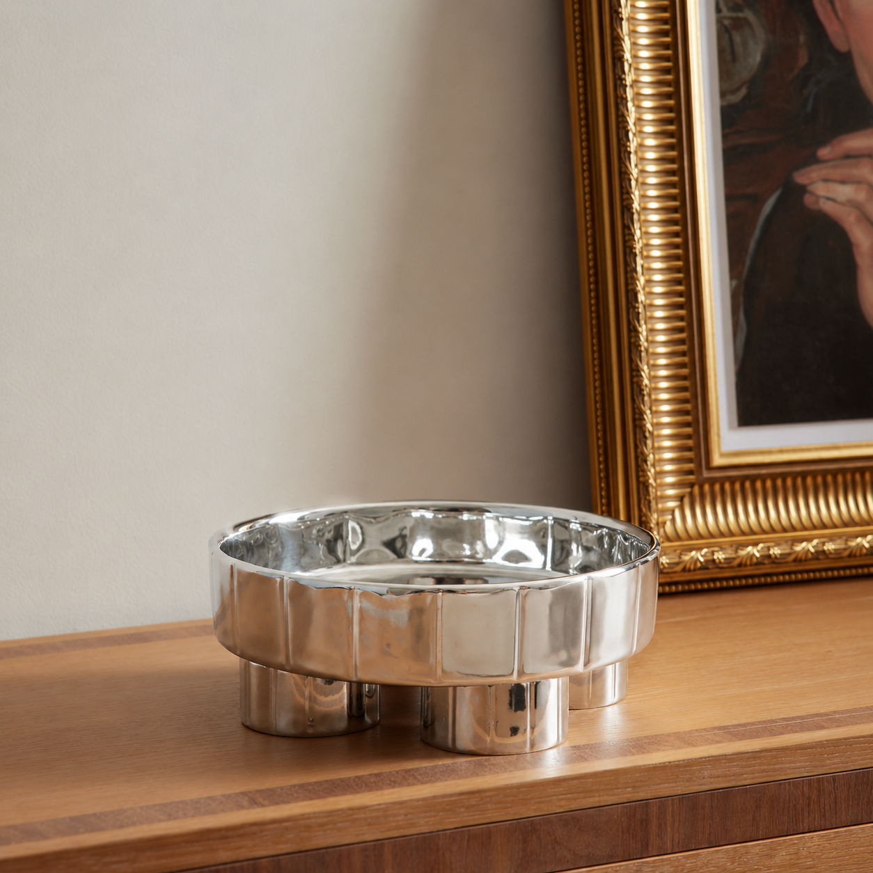 A polished silver metal ashtray with a fluted rim and three cylindrical feet, resting on a wooden console table next to a gold-framed picture.