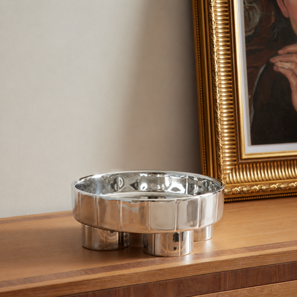 A polished silver metal ashtray with a fluted rim and three cylindrical feet, resting on a wooden console table next to a gold-framed picture.