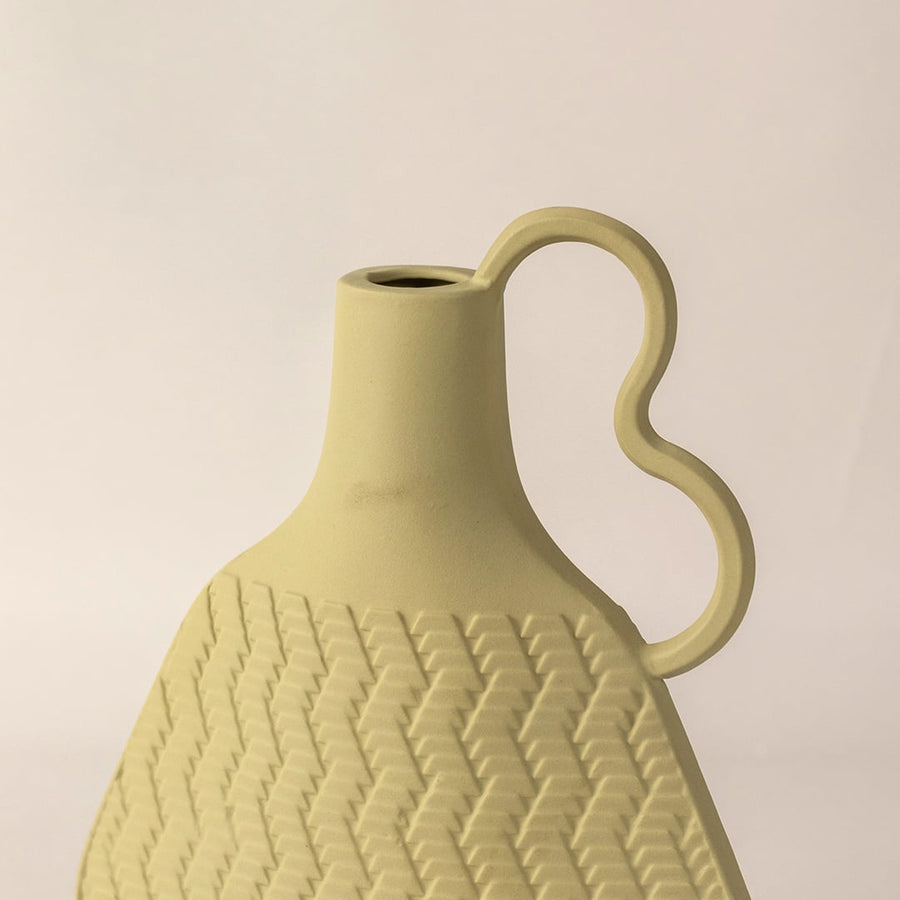 Light yellow ceramic vase with textured wave pattern and curved handle placed on white surface against neutral background showcasing modern artistic home decor styling.