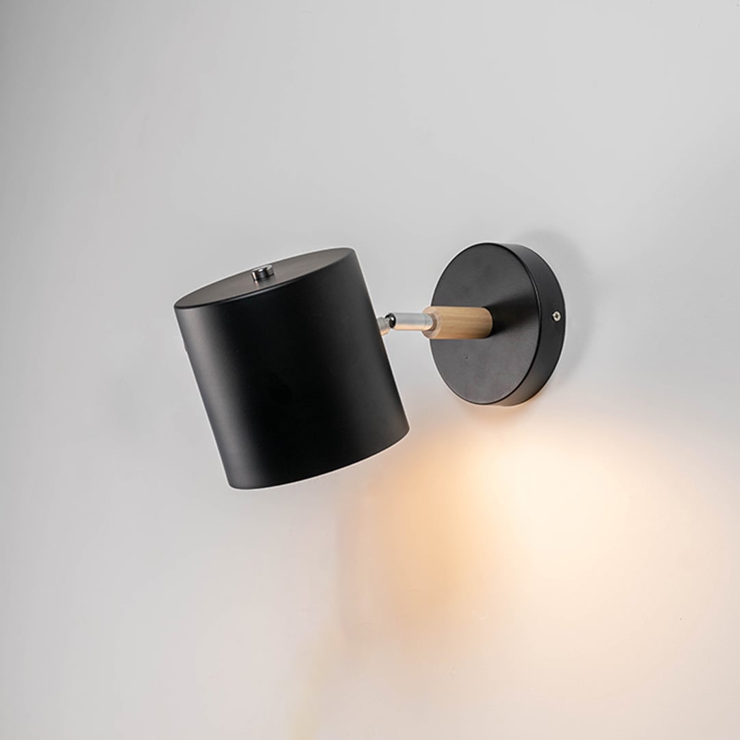 A black wall lamp with light on against a white background showing its cylindrical shade wooden connector and round base creating a clean modern illuminated look.