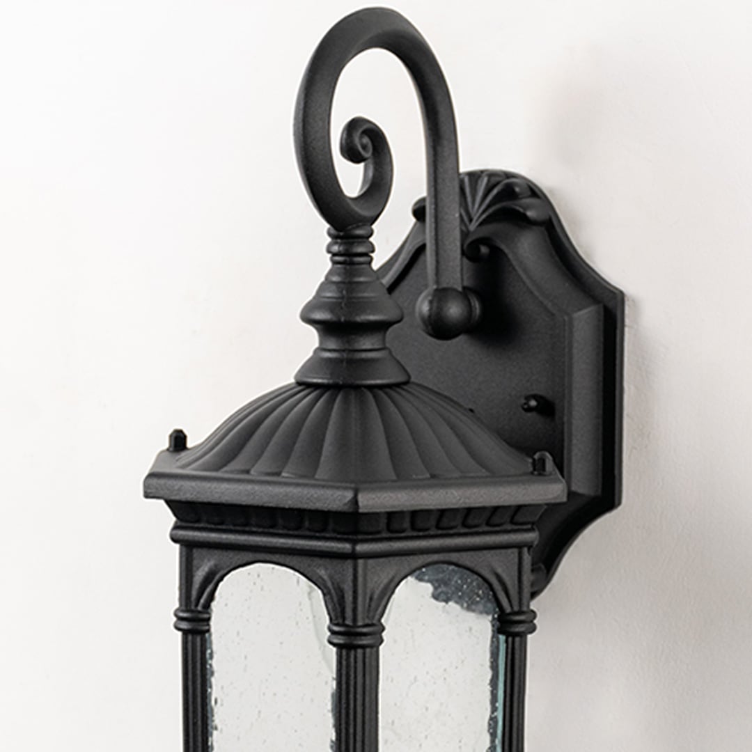 Closeup of outdoor wall lamp with clear textured glass and decorative black metal scroll arm is a vintage outdoor lighting fixture.