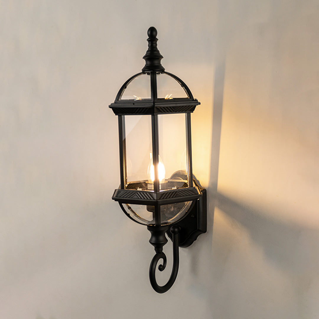 Black metal outdoor wall light on white background with an elongated glass lantern and bottom scroll for patio decor.