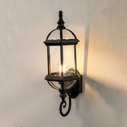 Black metal outdoor wall light on white background with an elongated glass lantern and bottom scroll for patio decor.