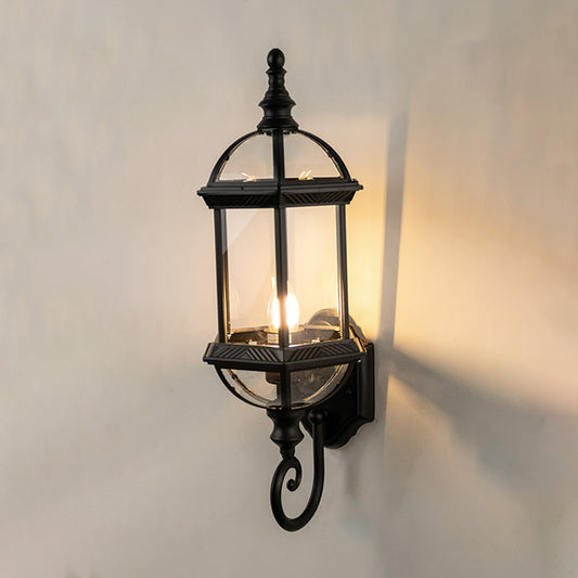 Black metal outdoor wall light on white background with an elongated glass lantern and bottom scroll for patio decor.