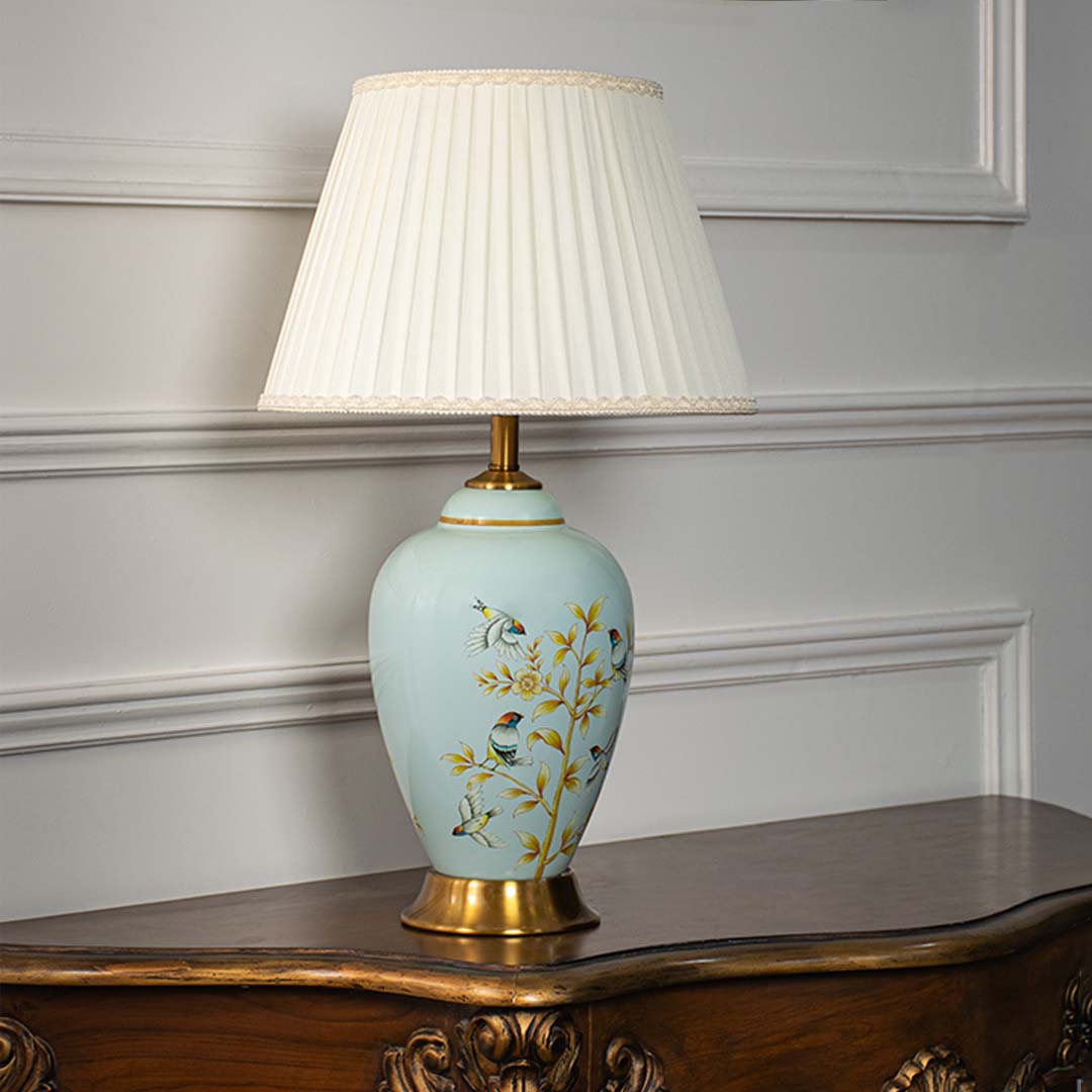Ceramic table lamp with pleated fabric shade blue base with floral and bird design and gold accents light off white background.