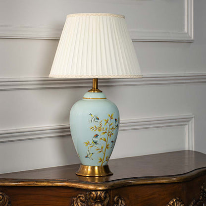 Ceramic table lamp with pleated fabric shade blue base with floral and bird design and gold accents light off white background.