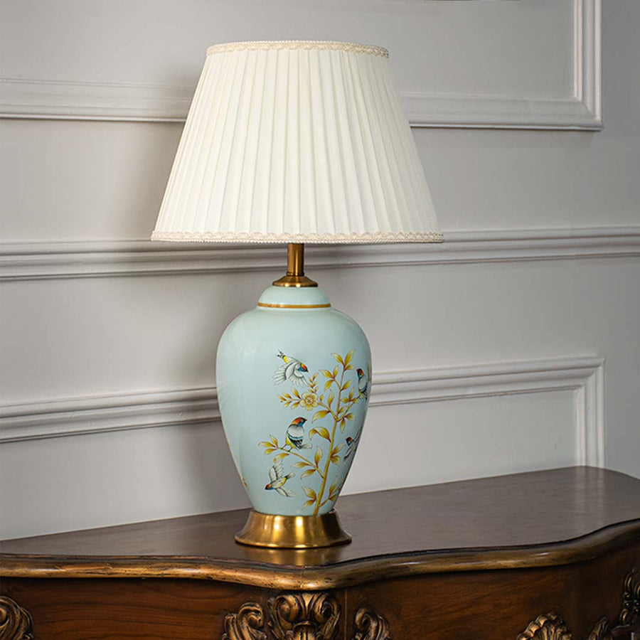 Ceramic table lamp with pleated fabric shade blue base with floral and bird design and gold accents light off white background.