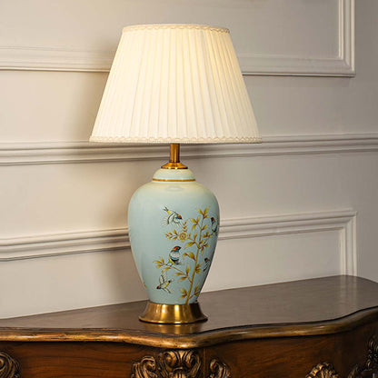 Ceramic table lamp with pleated shade light on white background blue floral and bird painted base with golden decorative stand.