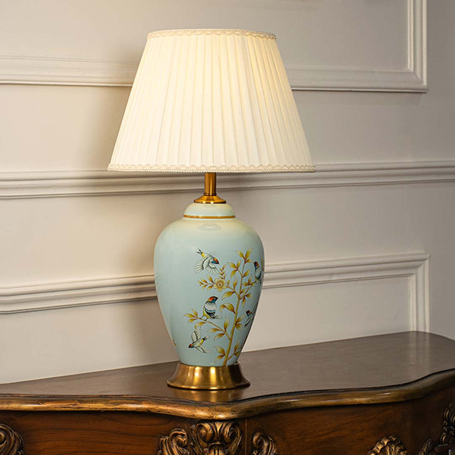 Ceramic table lamp with pleated shade light on white background blue floral and bird painted base with golden decorative stand.